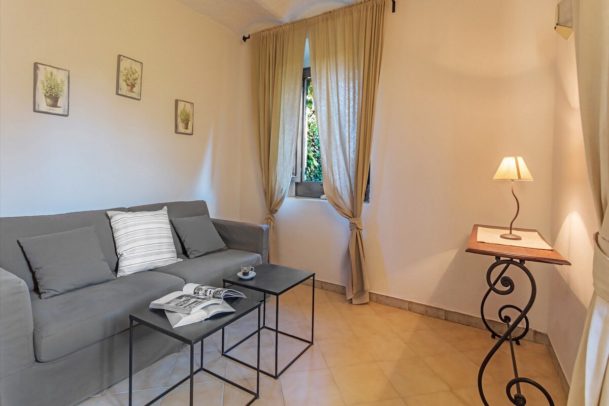 A living room with a grey sofa, a small coffee table with a magazine, a lamp on a side table, and a window with curtains.