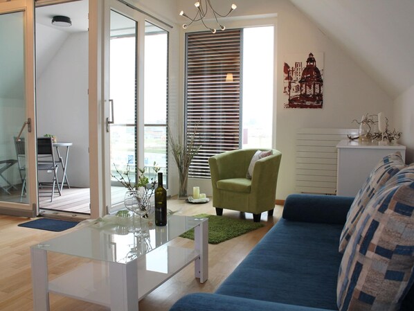 A modern living room with a glass coffee table, a blue sofa, a green armchair, and a dining area with a small table and chairs. - Charmante Ferienwohnung in Wismar am Hafen (Wismar)
