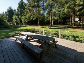 Um deck de madeira com mesas de piquenique, um parquinho com escorrega e uma área gramada com árvores.