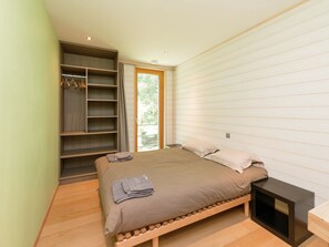 Quarto com cama, guarda-roupa de madeira, mesa de cabeceira e janela com vista para vegetação.