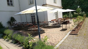 Un espace extérieur aménagé avec des tables et des chaises, entouré de plantes et bordé d’un bâtiment blanc.