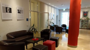 Un hall d’entrée avec des canapés en cuir, une table basse et un pouf rouge. Des affiches d’information sont accrochées au mur et une plante est placée au centre.