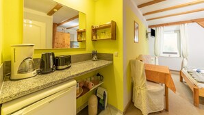 Cocina compacta con encimera de granito, cafetera, tostadora y microondas. Hay una mesa de madera con una silla y una cama con una ventana al fondo.