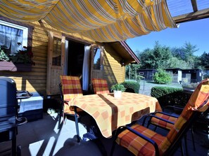 A wooden cabin with a patio area featuring a table and chairs, a grill, and a window with plants. - Bungalow in Klein Strtsmkendorf (Am Salzhaff)