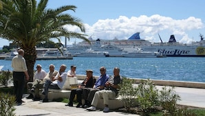 Un grupo de personas sentadas en un banco junto a la orilla, con un crucero y un barco pequeño al fondo.