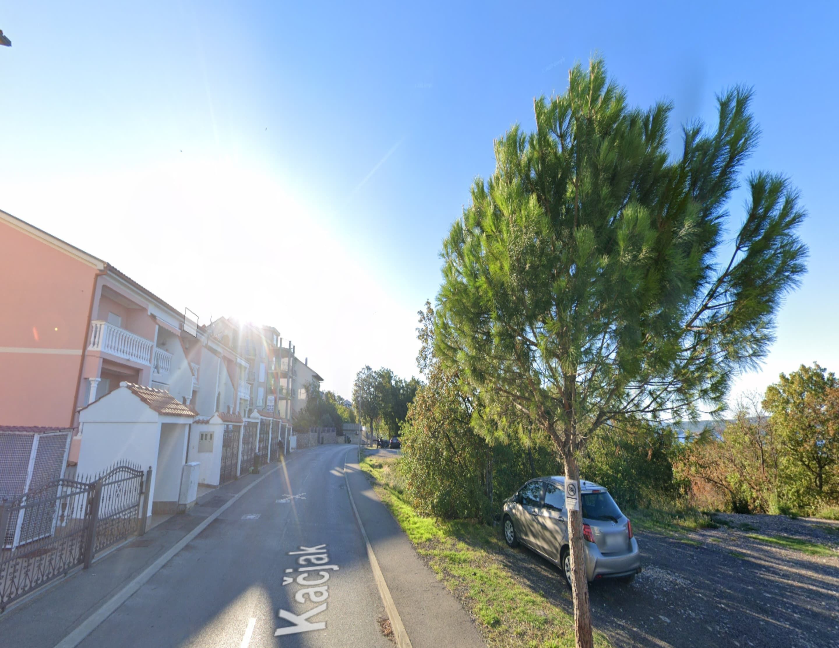 Una strada con un'auto parcheggiata sul lato, un albero e edifici sullo sfondo.