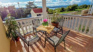 Varanda com mesa e cadeiras, plantas em vasos, vista para um corpo de água e montanhas.