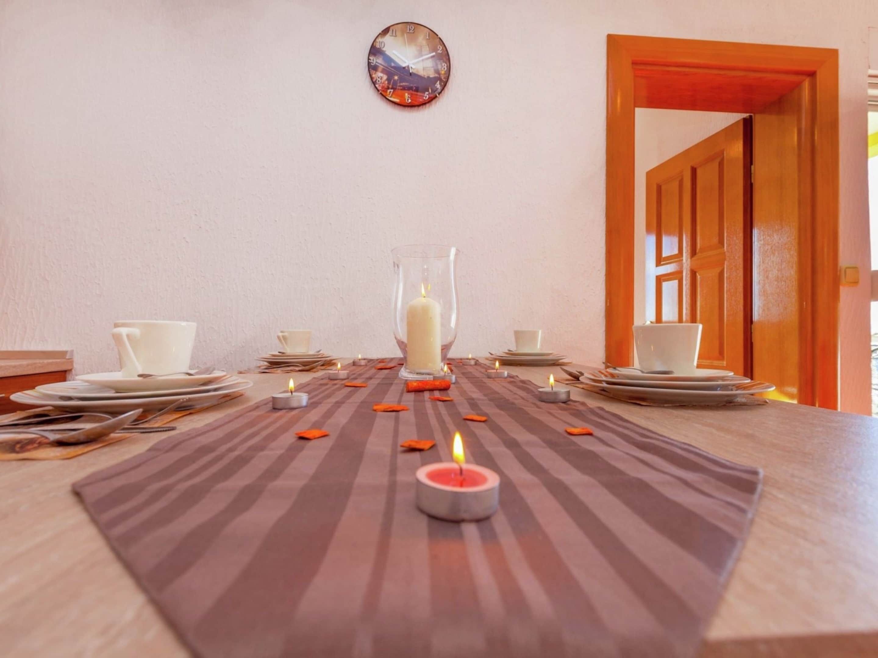 A dining table set with plates, cups, and candles, with a clock on the wall.