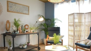 Een knus woongedeelte met een houten vloer, een witte tafel, een blauwe fauteuil, een zwarte metalen trolley met planten, en een groot raam met uitzicht.