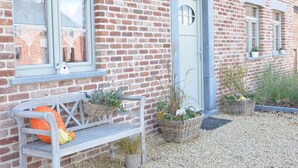 Een gebouw van baksteen met een witte deur, ramen en een bank met potplanten.