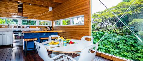 A modern kitchen with wooden walls, a dining area with white chairs, and a large window overlooking a lush garden.