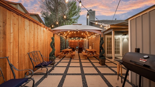 Un patio trasero con cerco de madera, mesas de picnic, una parrilla y luces colgantes.