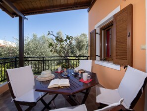 A wooden table with two white chairs, an open book, a mug, and a plate with food on a balcony with a view of trees and a building in the background. - Villa in Crete Near Beach and Olive Groves (Rethymno)
