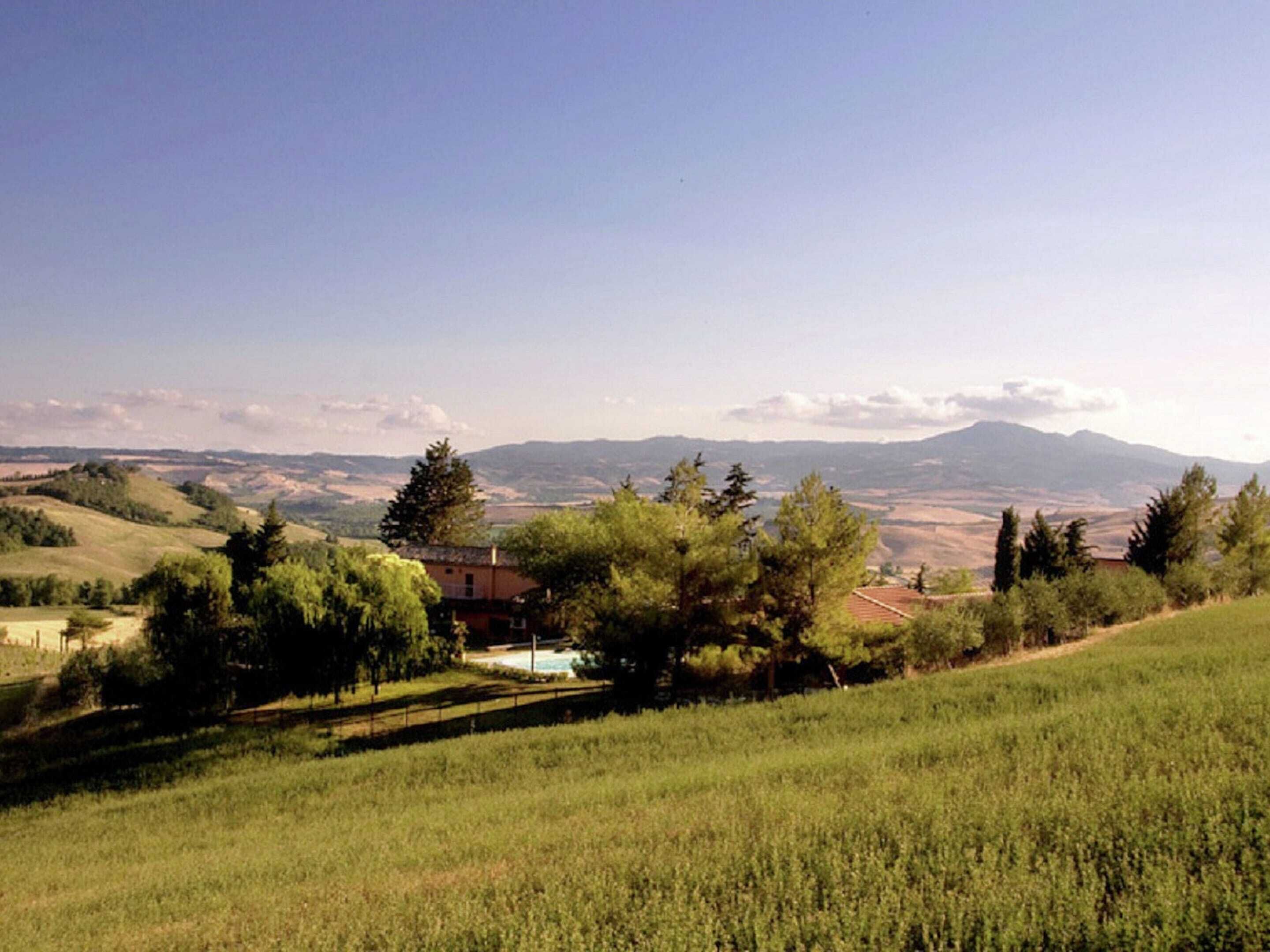 Un paysage rural avec des collines ondoyantes, une petite maison entourée d’arbres et, au loin, des montagnes sous un ciel dégagé.