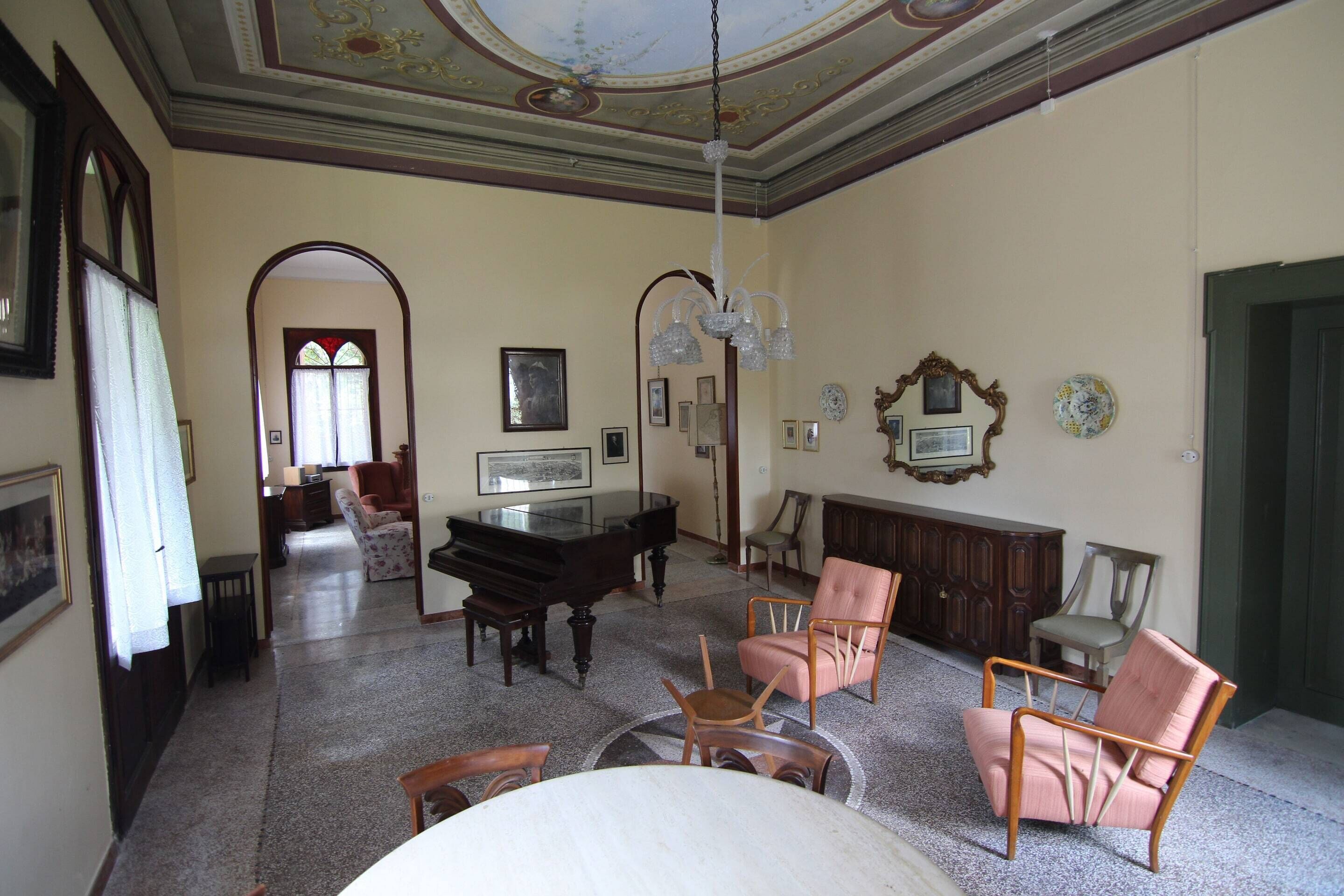 A spacious living room with a grand piano, multiple chairs, a dining table, and a chandelier.