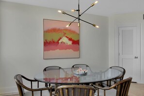 A dining area with a glass table surrounded by wicker chairs, a modern chandelier, and a large abstract painting on the wall.