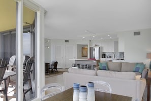 A modern living room with a sofa, dining table, and chairs. There is a kitchen area with a refrigerator and bar stools.