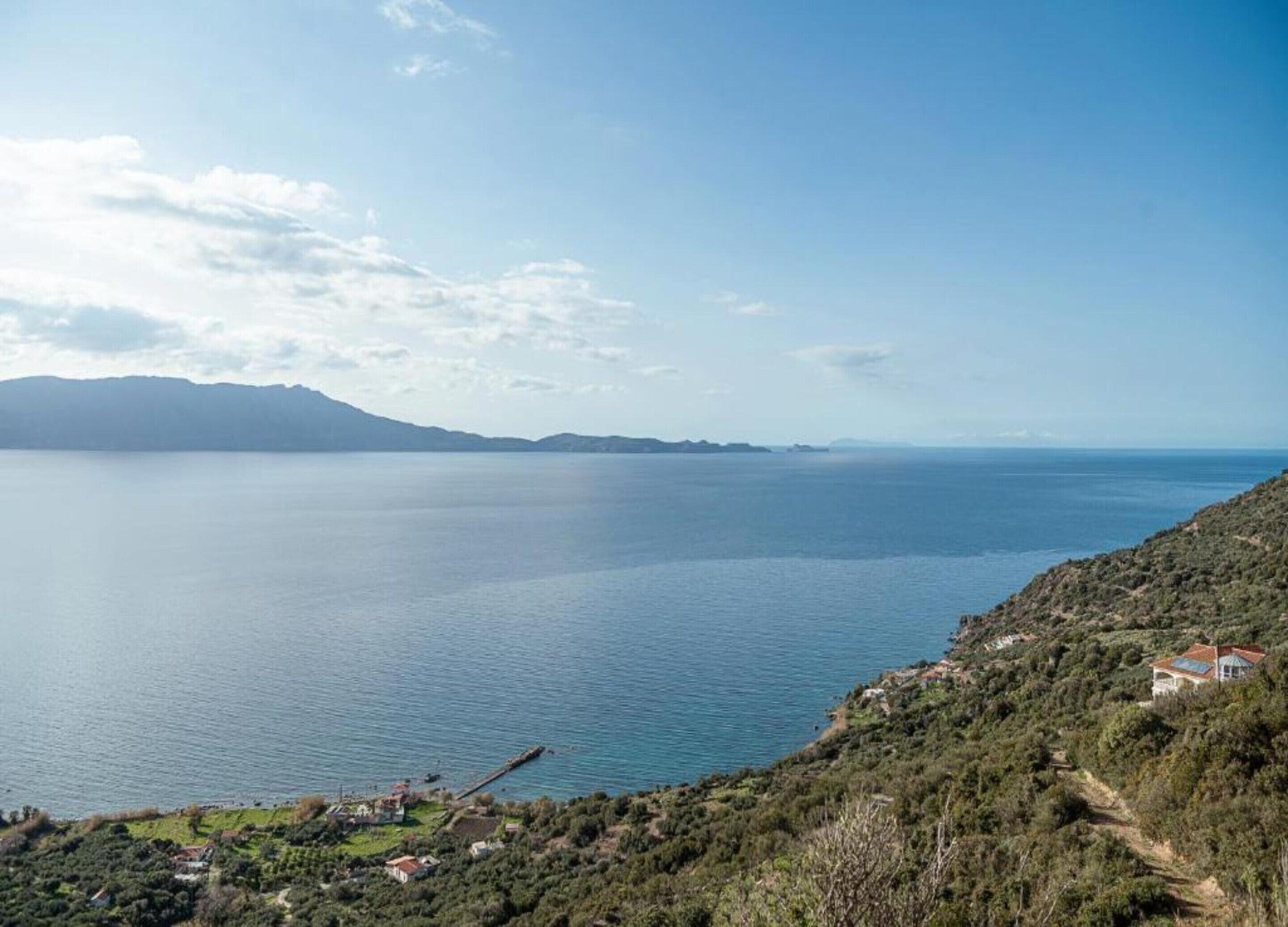 A coastal landscape with a clear view of the sea, a distant mountain range, and a few buildings on the hillside.