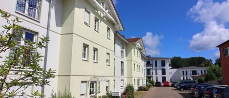 A row of light-colored buildings with a paved walkway and parked cars.