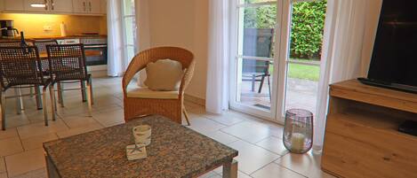 A modern kitchen with white cabinets, a dining area with a table and chairs, a wicker chair, and a television on a wooden stand.
