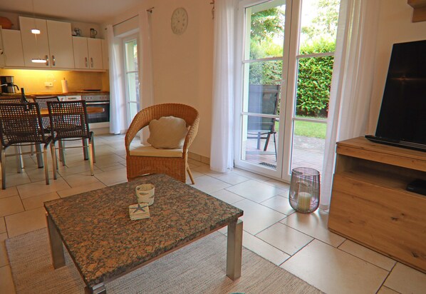 A modern kitchen with white cabinets, a dining area with a table and chairs, a wicker chair, and a television on a wooden stand. - Moderne Ferienwohnung in Graal-muritz, Strandnah (Graal Mueritz)