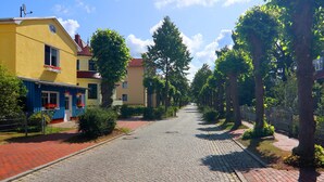 Eine Kopfsteinpflasterstraße, gesäumt von Bäumen und Gebäuden, darunter ein gelbes Gebäude mit einer blauen Markise und einem roten Dach.