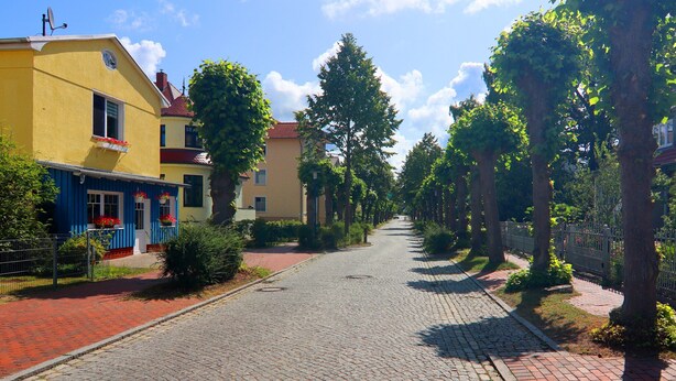 A cobblestone street lined with trees and buildings, including a yellow building with a blue awning and a red roof. - Moderne Ferienwohnung in Graal-muritz, Strandnah (Graal Mueritz)