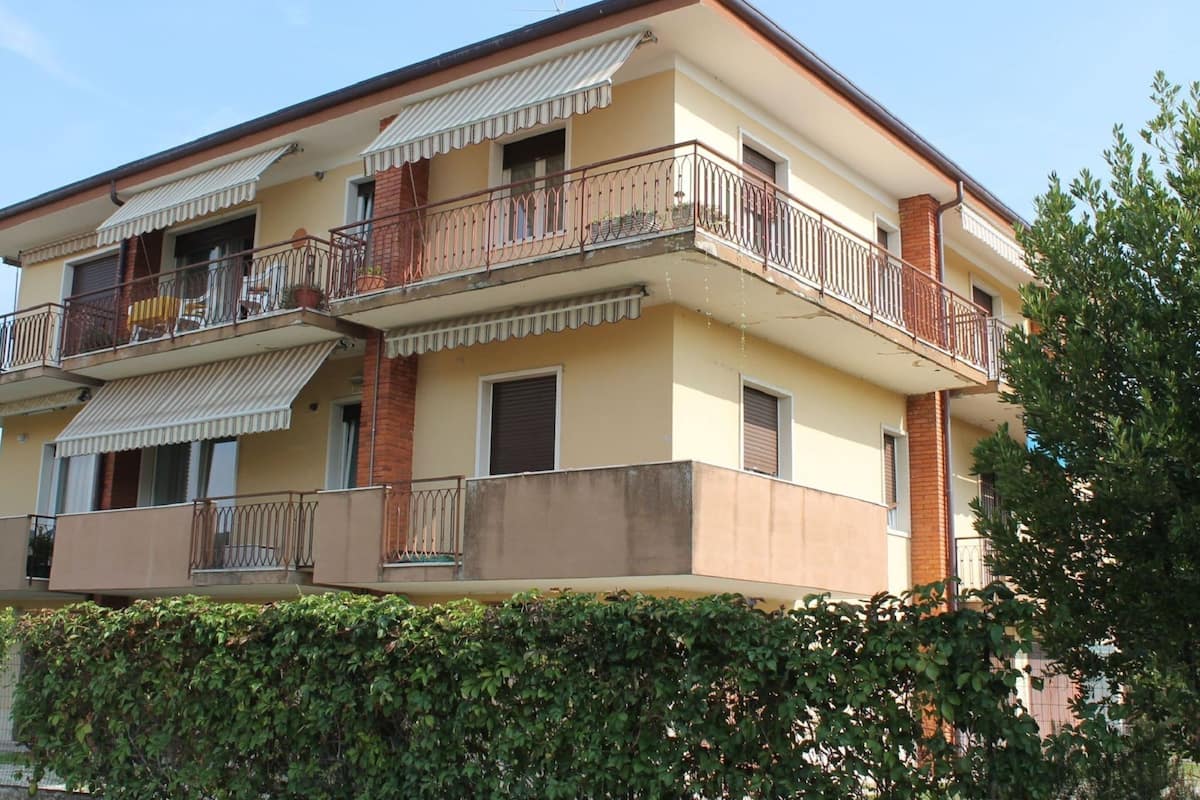 A multi-story residential building with balconies, a flat roof, and a hedge in front.