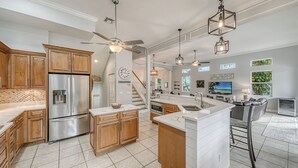 A modern kitchen with a stainless steel refrigerator, wooden cabinets, and a large island with a sink. There are pendant lights, a ceiling fan, and a view of the living area with a TV and seating. - Siesta Soul | Multilevel Home Located in Siesta Key Village w/ Private Pool, Fire Pit, Walk to Beach (Siesta Key)