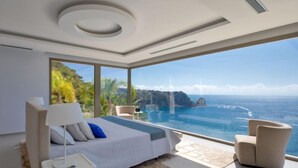 A modern bedroom with a large bed, a bedside table, a chair, and a view of the ocean and cliffs. - Emma on the Roc by Interhome (Jávea)