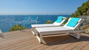 A wooden deck with two white sun loungers, each with a blue towel, overlooking a clear blue sea and a distant coastline. - Emma on the Roc by Interhome (Jávea)