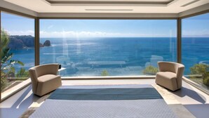 A modern room with a large panoramic window offering a view of the ocean and cliffs. There are two beige armchairs and a blue rug on the floor. - Emma on the Roc by Interhome (Jávea)