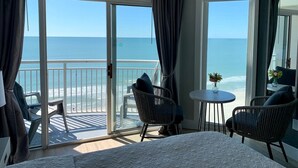 A room with a bed, two chairs, a small table, and a view of the ocean through large windows.