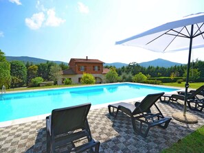A swimming pool with lounge chairs and an umbrella, surrounded by a house and mountains.