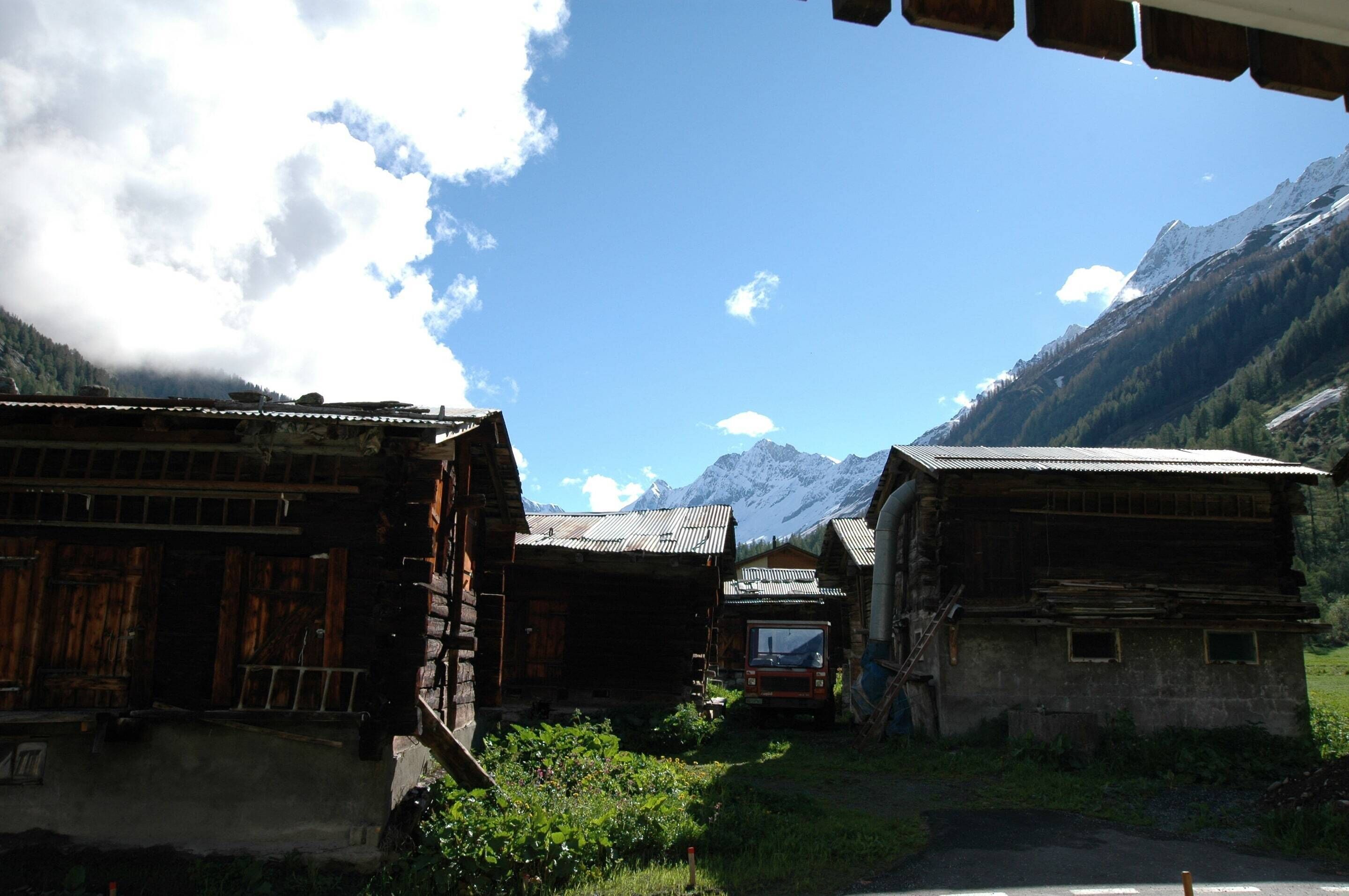 A rustic mountain village with wooden buildings, a dirt road, and snow-capped mountains in the background.