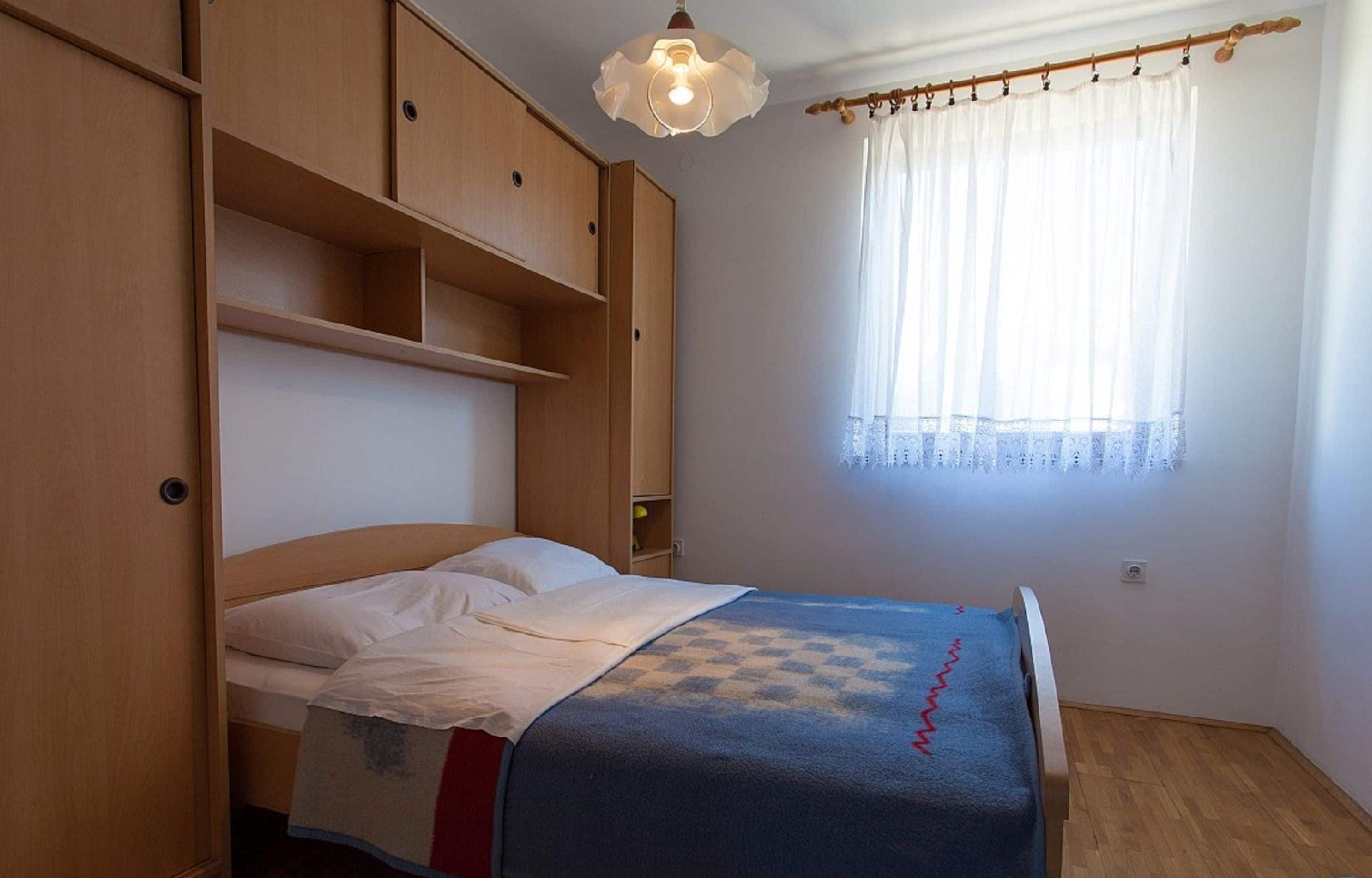 Ein Schlafzimmer mit Bett, Holzschrank und Fenster mit durchsichtigen Vorhängen.