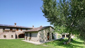 Un bâtiment en pierre avec un toit en tuiles, une cour verdoyante et un arbre qui offre de l’ombre.