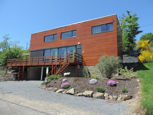 A modern two-story building with a wooden deck, stone foundation, and a landscaped garden with rocks and flowers. - Lovely Modern Cottage With Sauna, Bubble Baths and Even a Beach on the River (Stavelot)