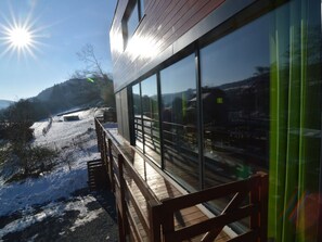 Ein modernes Gebäude mit großen Fenstern, einer Holzterrasse und einer verschneiten Landschaft im Hintergrund.