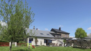 A two-story house with a sloped roof, a stone facade, and a large front yard with a play area. - Cushy Holiday Home With Fenced Garden (Tenneville)