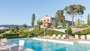 Piscina com espreguiçadeiras e guarda-sóis, cercada por vegetação e um edifício de pedra ao fundo.