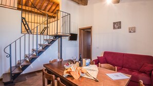 Uma sala de jantar com mesa de madeira, cadeiras e um sofá vermelho. Uma escadaria com grades de metal é visível ao fundo.