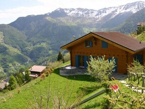 A wooden house with a sloped roof, surrounded by lush green hills and snow-capped mountains in the background. - Chalet Alpina (Riddes)