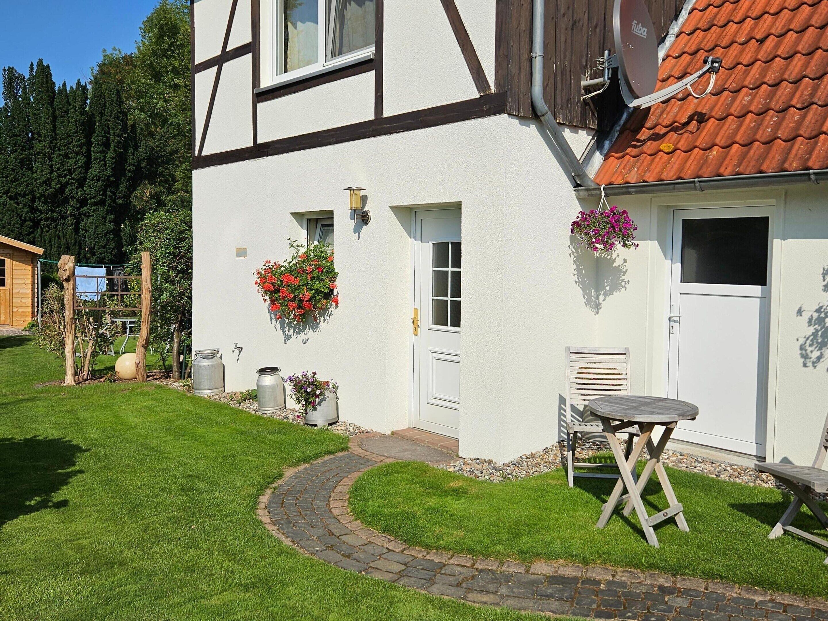 Un bâtiment blanc surmonté d’un toit recouvert de tuiles rouges, un petit jardin avec un chemin dallé et du mobilier d’extérieur.