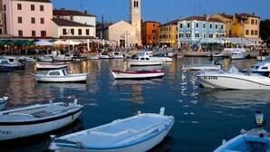 A marina with various boats docked, a prominent church tower, and colorful buildings along the waterfront.