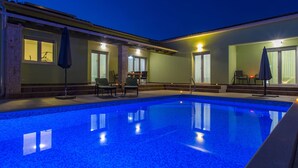 A swimming pool with two chairs and a table, surrounded by a building with large windows and a covered patio area.