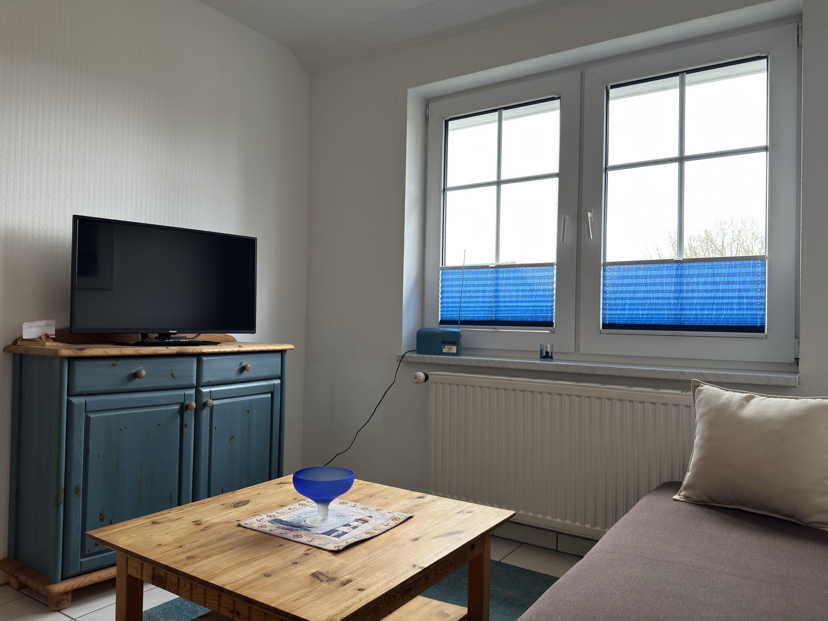 Ein Zimmer mit Fernseher auf einer Holzkommode, einem Tisch mit blauer Schüssel und einem Fenster mit Jalousien.