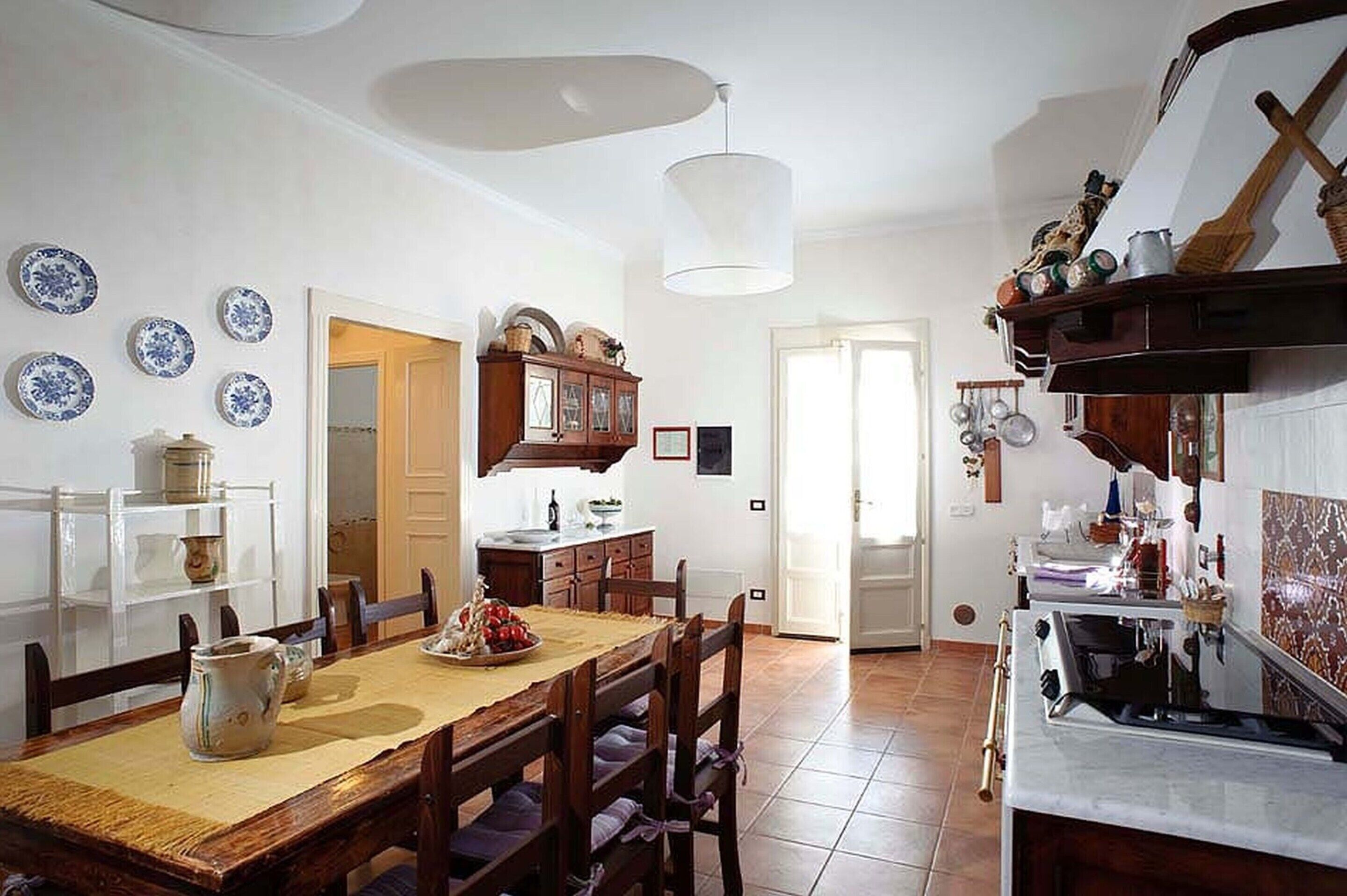 A dining area with a wooden table and chairs, a hanging light fixture, and a kitchen area with a stove and utensils.
