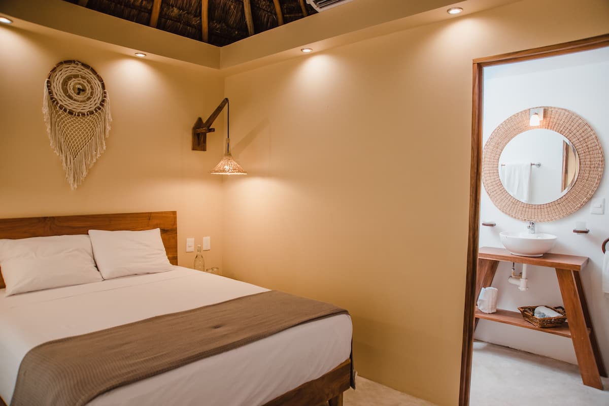 A bedroom with a bed, bedside lamp, wall art, and a bathroom with a round mirror, sink, and shelf.