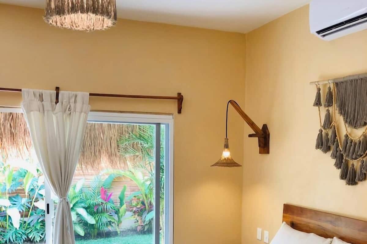 A bedroom with a large bed, a bedside lamp, a ceiling fan, and a view of greenery outside.